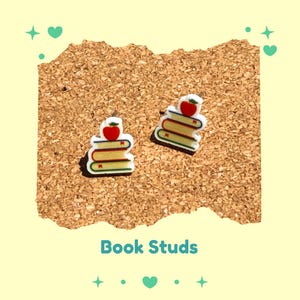 Può includere: Orecchini a forma di libri con una mela rossa. Gli orecchini sono bianchi con dettagli verdi, gialli e rossi. Lo sfondo è una bacheca di sughero con la scritta "Book Studs".