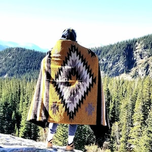 May include: A person wearing a yellow, black, and white patterned blanket stands on a rocky outcrop overlooking a forest of evergreen trees.