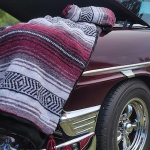 May include: A maroon and white classic car with the hood open, revealing a red and white striped blanket inside. The car is parked on a grassy lawn.