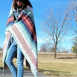 May include: A colorful woven blanket with a striped pattern in shades of red, blue, and green, with a geometric design on the edges. The blanket has tassels on the corners. The person is wearing blue jeans and a denim jacket.