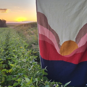 May include: A woven tapestry with a sunset design featuring a large orange sun, pink, red, and brown mountains, and a dark blue sky. The tapestry is hanging in a field of green plants.