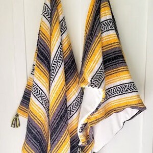 May include: Two hanging serape blankets with yellow, black, white, and gray stripes and geometric patterns. Each blanket has a tassel at the bottom. The blankets are hanging on a white wall.