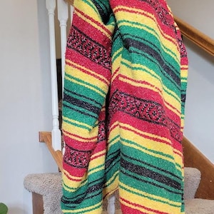 May include: A woven poncho featuring horizontal stripes in vibrant red, yellow, green, and black. The garment includes intricate geometric patterns, a solid black interior lining, and decorative yarn tassels at the bottom corners with a small white label.