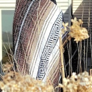 May include: A woven blanket featuring a geometric pattern in shades of brown, white, and black. The textile displays a repeating diamond motif and striped borders, draped over the shoulders of a person wearing dark sunglasses outdoors.