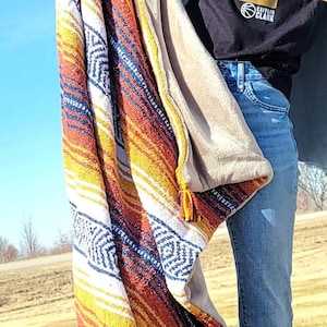 May include: A rectangular throw blanket featuring a vibrant, multi-colored striped pattern in shades of orange, yellow, white, and blue with geometric diamond motifs. The blanket has a soft, solid-colored lining and decorative tassels at the corners.