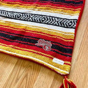 May include: A woven blanket featuring horizontal stripes in red, yellow, white, and black. The blanket includes a red border, a red and yellow tassel at the corner, a San Francisco 49ers logo patch, and a small white tag labeled House of Armor.