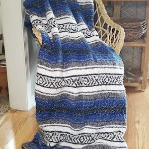 May include: A woven blanket featuring a repeating pattern of horizontal stripes in shades of royal blue, white, black, and gray. The textile has a textured appearance with decorative fringe tassels at the corners and a small white label on the edge.