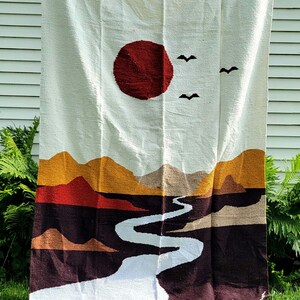 May include: A woven tapestry featuring a landscape design. The tapestry depicts a red sun, two black birds, and a winding white river. The landscape includes mountains in shades of brown, orange, and red. The background is white.