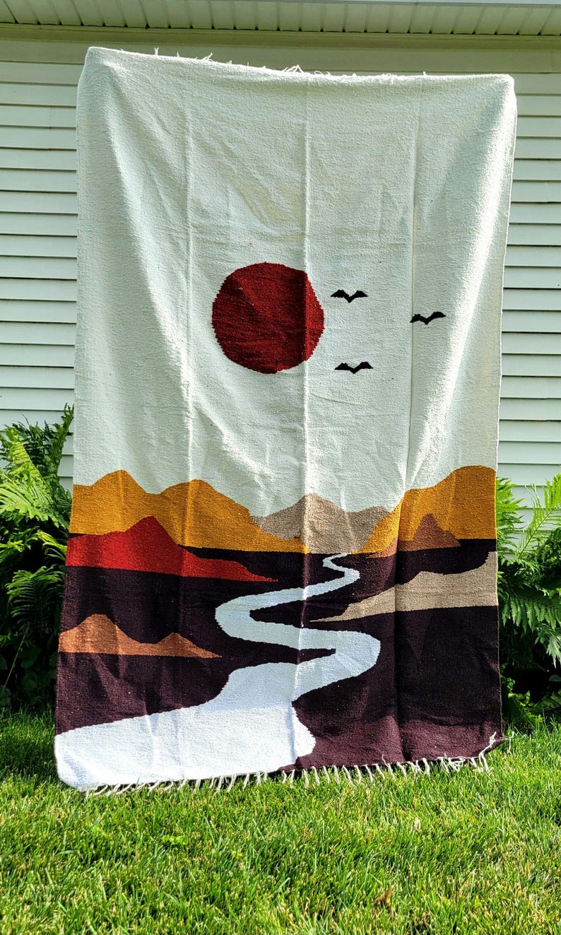 May include: A woven tapestry featuring a landscape design. The tapestry depicts a red sun, two black birds, and a winding white river. The landscape includes mountains in shades of brown, orange, and red. The background is white.