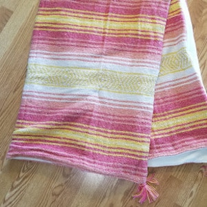 May include: A pink and yellow striped throw blanket with a fringe on one end. The blanket is folded and lying on a wooden floor.