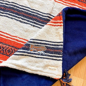 May include: A woven blanket featuring a striped pattern in orange, cream, and navy blue. The blanket has a solid navy blue fleece backing and a Denver Broncos logo patch. A small tag reads House of Armor, and a decorative tassel hangs from the corner.