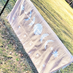 May include: A rectangular beige woven blanket featuring a repeating white graphic print of the lunar cycle, including a full moon, crescent moons, and various star shapes, with fringed edges along the shorter sides of the textile fabric.
