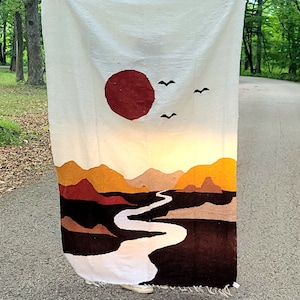 May include: A white woven blanket with a landscape design. The design features a large red sun, mountains in shades of brown and yellow, a winding river, and two birds. The blanket is fringed at the bottom.