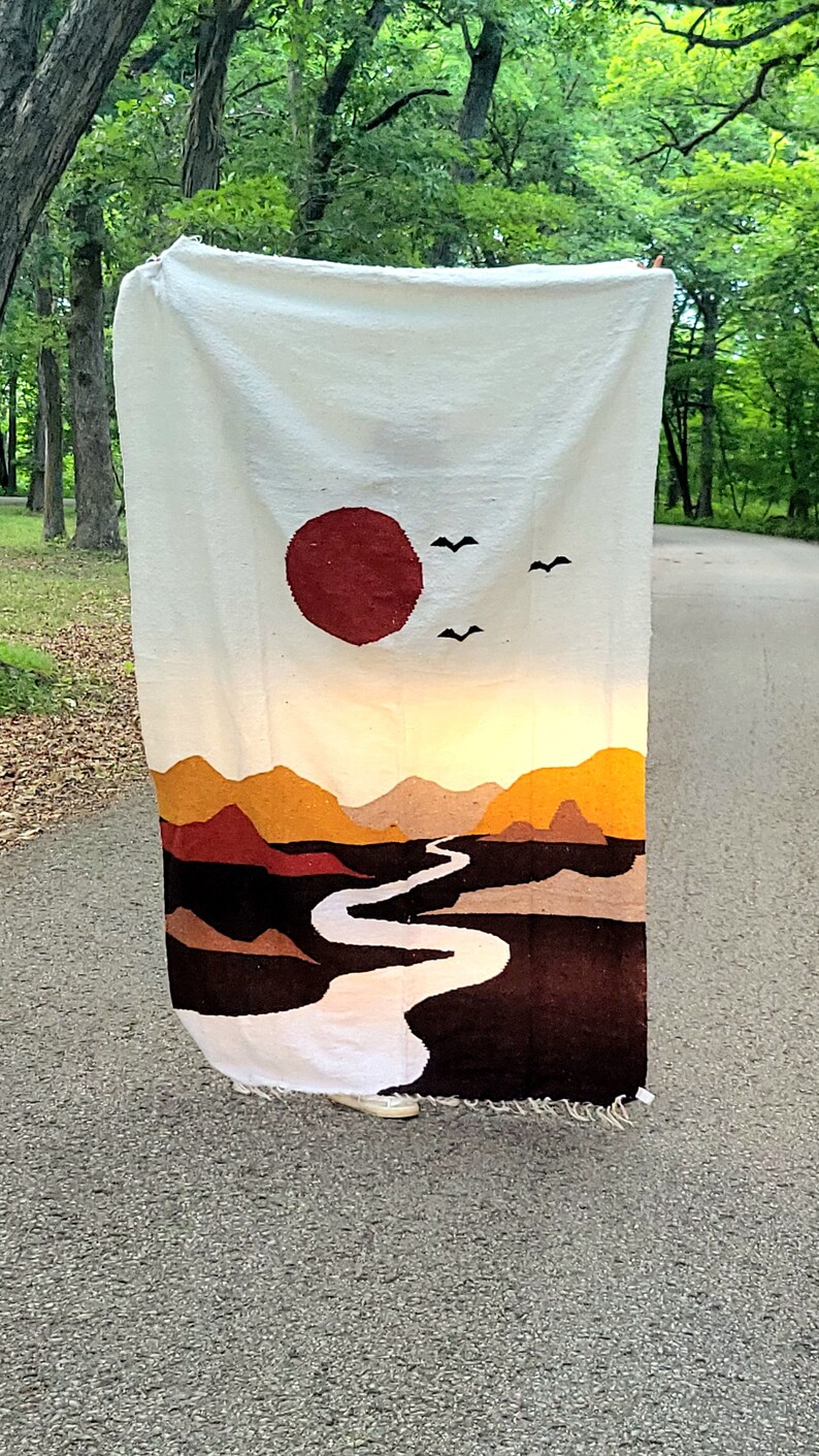 May include: A white woven blanket with a landscape design. The design features a large red sun, mountains in shades of brown and yellow, a winding river, and two birds. The blanket is fringed at the bottom.