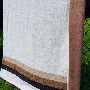 May include: A rectangular textile blanket featuring a large off-white central panel. One side edge is finished with a wide, solid brown border. The bottom section displays horizontal stripes in shades of light tan, medium brown, and dark brown.