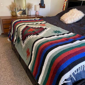 May include: A woven blanket featuring a central diamond pattern in red, green, and black, surrounded by horizontal stripes in blue, red, green, and white. The blanket has white fringe along the edges and is draped over a dark bed frame.