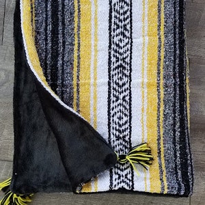 May include: A folded blanket with a striped pattern in black, yellow, white, and gray. The blanket has a black, plush underside and yellow tassels. The design features a central geometric pattern.