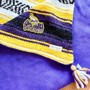 May include: A purple fleece blanket featuring a folded corner with a woven striped pattern in purple, yellow, and white. The corner displays a Minnesota Vikings logo patch and a small white tag reading House of Armor. A multi-colored tassel is attached.