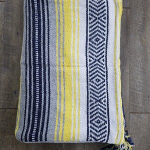 May include: Folded woven blanket with a striped pattern in navy blue, yellow, and gray. The blanket features a geometric design on one side and a tassel trim. The blanket is made of a soft, textured material.
