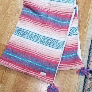 May include: A folded woven blanket featuring horizontal stripes in shades of pink, white, and teal. The fabric displays a repeating geometric diamond pattern across the center and ends with decorative multi-colored tassels in pink and teal.