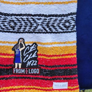 May include: A woven blanket featuring horizontal stripes in red, yellow, white, and black. An embroidered patch depicts a basketball player in a blue jersey with the number 22, text reading "CZ CHK #22 FROM LOGO", and a small white tag labeled "HOUSE OF ARMOR".