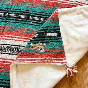 May include: A woven blanket featuring horizontal stripes in white, orange, and teal with black accents. The fabric has a soft white underside and a corner tassel. An embroidered Miami Dolphins logo is visible on the patterned side of the blanket.