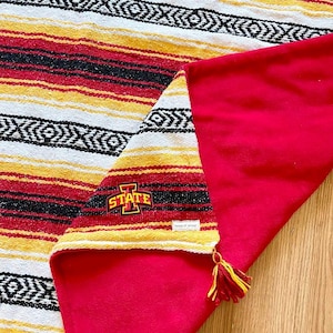 May include: A woven blanket featuring horizontal stripes in red, yellow, white, and black with a geometric pattern. One corner is folded back to reveal a solid red underside and a small tassel. An Iowa State University logo is embroidered on the corner.