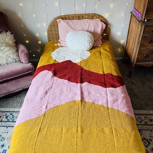 May include: A twin-sized bed featuring a woven blanket with a large-scale abstract pattern in mustard yellow, light pink, and deep red. The blanket has a textured surface and a small white rectangular tag at the bottom right corner with black text.