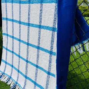 May include: A rectangular textile throw featuring a white base with a grid pattern of bright blue lines. The fabric has a textured weave and is finished with white fringe along the bottom edge. A solid deep blue panel runs along the right side.