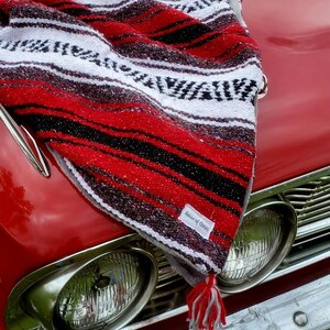 May include: A red, black, and white striped serape blanket with a tassel, draped over the hood of a classic red car. The blanket has a tag that reads "House of Canna".