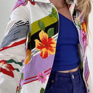 Op de afbeelding: Een wit jack met rits met een kleurrijke bloemenprint. Het jack heeft een gestreept patroon in paars, groen en zwart met rode accenten. De bloemen zijn geel, rood, roze en oranje.