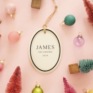 May include: An oval Christmas ornament with the name "JAMES" and "FIRST CHRISTMAS 2024" printed on it. The ornament is surrounded by small, colorful Christmas tree decorations and glass ball ornaments on a pink background.