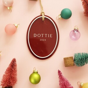 May include: A festive arrangement on a pink background featuring Christmas ornaments in various colors, miniature trees, and a burgundy oval tag with the name "DOTTIE" and the year 2024.