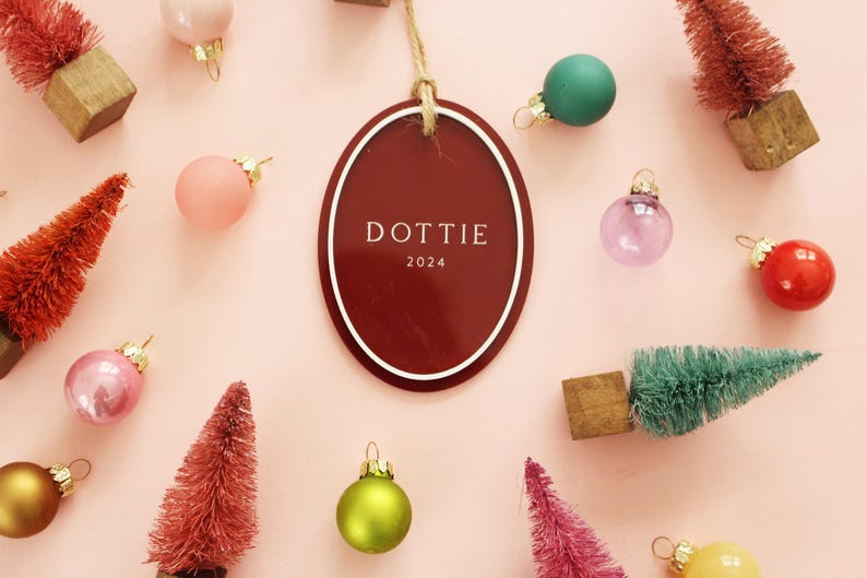 May include: A festive arrangement on a pink background featuring Christmas ornaments in various colors, miniature trees, and a burgundy oval tag with the name "DOTTIE" and the year 2024.