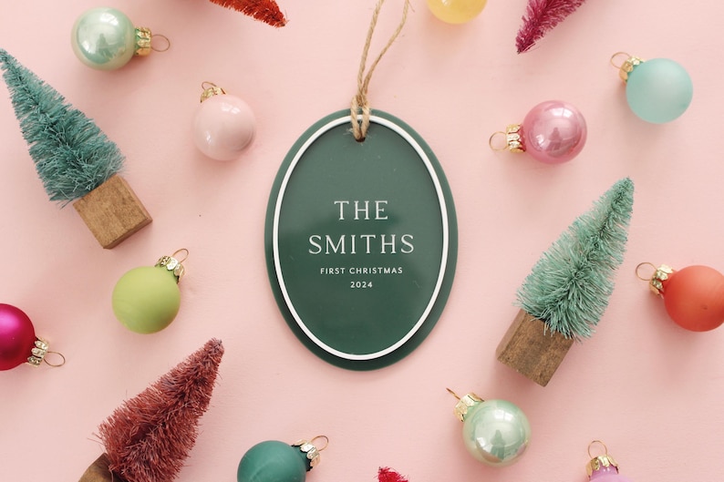 May include: A festive Christmas ornament with the text "THE SMITHS FIRST CHRISTMAS 2024" in white on a green oval. The ornament is surrounded by miniature Christmas trees and colorful ornaments in shades of pink, green, and red.