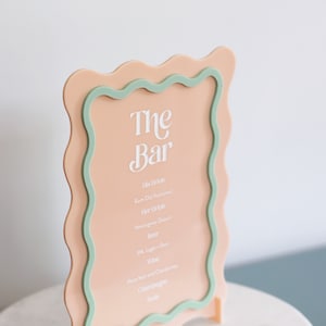 May include: A peach-colored acrylic sign with a wavy green border. The sign reads "The Bar" and lists various drink options, including rum old fashioned, hot drinks, beer, wine, and champagne.