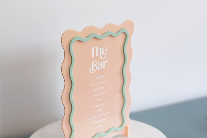 May include: A peach-colored acrylic sign with a wavy green border. The sign reads "The Bar" and lists various drink options, including rum old fashioned, hot drinks, beer, wine, and champagne.