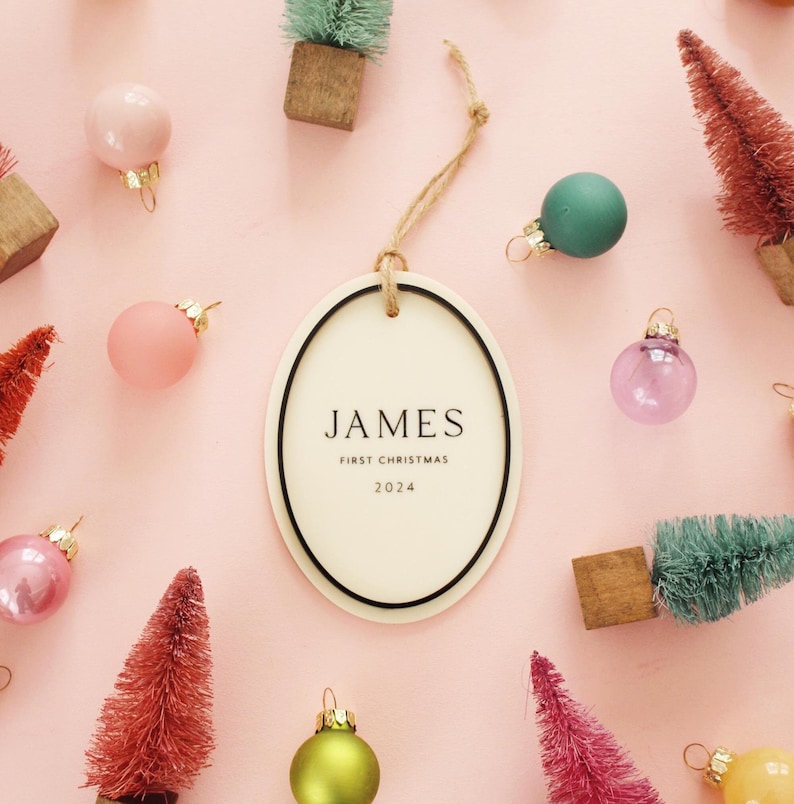 May include: An oval Christmas ornament with the name "JAMES" and "FIRST CHRISTMAS 2024" printed on it. The ornament is surrounded by small, colorful Christmas tree decorations and glass ball ornaments on a pink background.