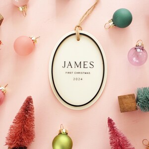 May include: Oval Christmas ornament with the name "JAMES" and the text "FIRST CHRISTMAS 2024". The ornament is surrounded by miniature Christmas trees and colorful ornaments on a pink background.