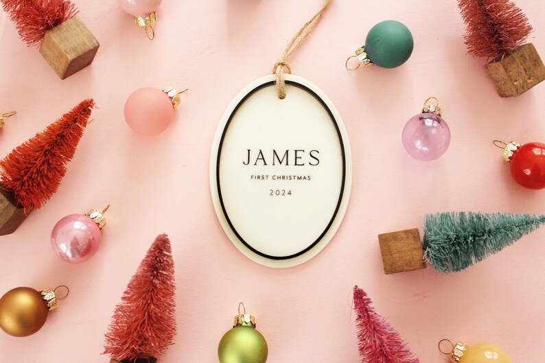 May include: Oval Christmas ornament with the name "JAMES" and the text "FIRST CHRISTMAS 2024". The ornament is surrounded by miniature Christmas trees and colorful ornaments on a pink background.