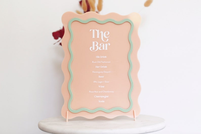 May include: A pink acrylic sign with a green border and wavy edges. The sign reads "The Bar" and lists drink options: His Drink, Ram Old Fashioned, Her Drink, Hemingway Daiquiri, Beer, Wine, Post Wine and Chardonnay, Champagne, Soda.