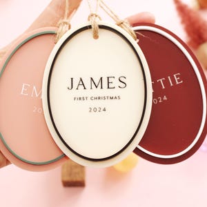 May include: Three oval-shaped Christmas ornaments in shades of pink, cream, and burgundy, each with a white border and a jute string for hanging. The center ornament reads "JAMES FIRST CHRISTMAS 2024". The other ornaments have names and the year 2024.