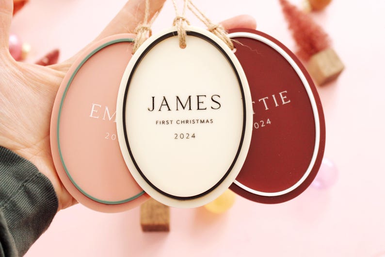 May include: Three oval-shaped Christmas ornaments in shades of pink, cream, and burgundy, each with a white border and a jute string for hanging. The center ornament reads "JAMES FIRST CHRISTMAS 2024". The other ornaments have names and the year 2024.