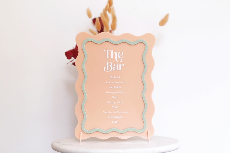 May include: A peach-colored sign with a wavy border and a green inner border. The sign says "The Bar" in white text and lists drink options: His Drinks, Her Drinks, Beer, Wine, Champagne, Soda.