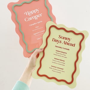 May include: Two colorful signs with wavy edges. The pink sign says "Happy Camper" and lists "Campfire Spirits", "Wine", "Signature Cocktail", and "Water". The yellow sign says "Sonny Days Ahead" and lists "Specialty Cocktails", "Sunburn Margarita", "Starburst Mimosa", and "Summer Lemonade".