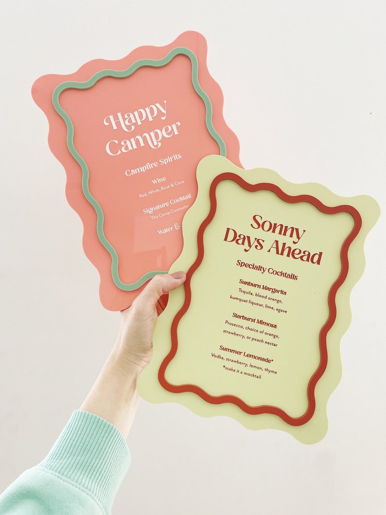 May include: Two colorful signs with wavy edges. The pink sign says "Happy Camper" and lists "Campfire Spirits", "Wine", "Signature Cocktail", and "Water". The yellow sign says "Sonny Days Ahead" and lists "Specialty Cocktails", "Sunburn Margarita", "Starburst Mimosa", and "Summer Lemonade".