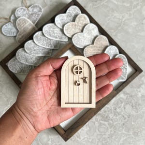 May include: A small, unfinished wooden fairy door with a round window and a keyhole is held in a hand. The door is set against a backdrop of felt hearts and a wooden frame, suggesting a craft or decorative item.