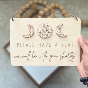 May include: Wooden sign with a light wood finish, featuring three decorative moon phases and botanical details. The sign reads "PLEASE HAVE A SEAT, we will be with you shortly" in a script font. A leather cord is attached for hanging.