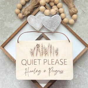 May include: A wooden sign with the words "QUIET PLEASE" and "Healing in Progress" engraved on it. The sign features a floral design and is hanging by a brown leather cord. Two wooden hearts and wooden beads are also visible.