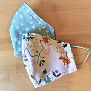 May include: Two fabric face masks. One is light blue with white paw prints. The other is white with a jungle animal print featuring a sloth, toucan, flamingo, and snakes.